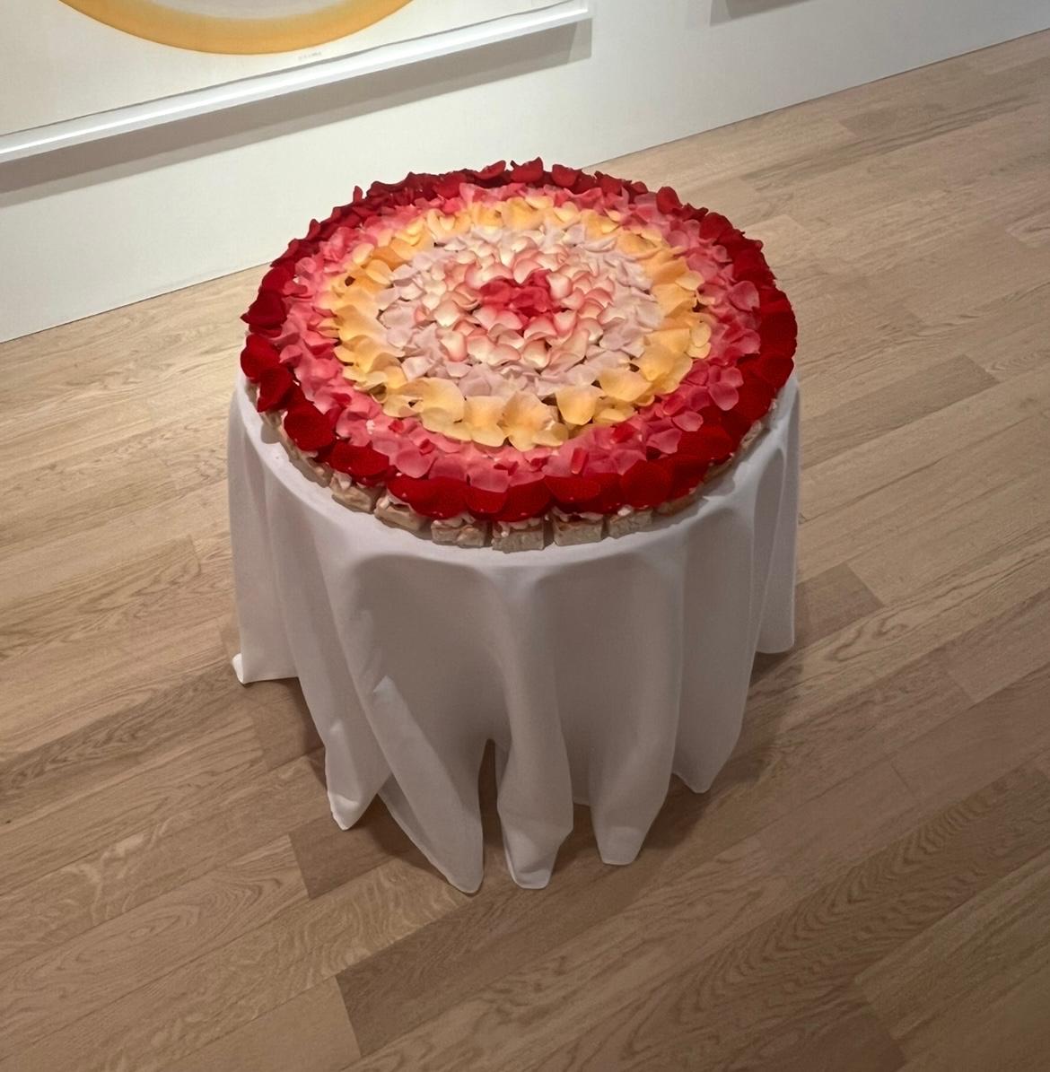 A round floral arrangement made of layered rose petals in red, pink, and yellow tones, displayed on a white draped tablecloth in an indoor room with light wood flooring.